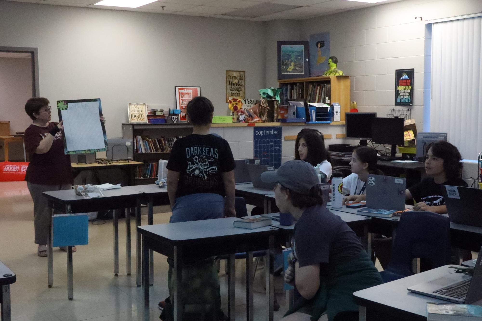 Mrs. Brady explains a new feature in the Battle of the Books club. When a student finishes a book, they put a checkmark on the chart next to their name.